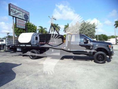 Ford F-600 rollback tow truck with a flatbed, black cab, and white mid-section, side view showing long bed and equipment; dealership lot in background.