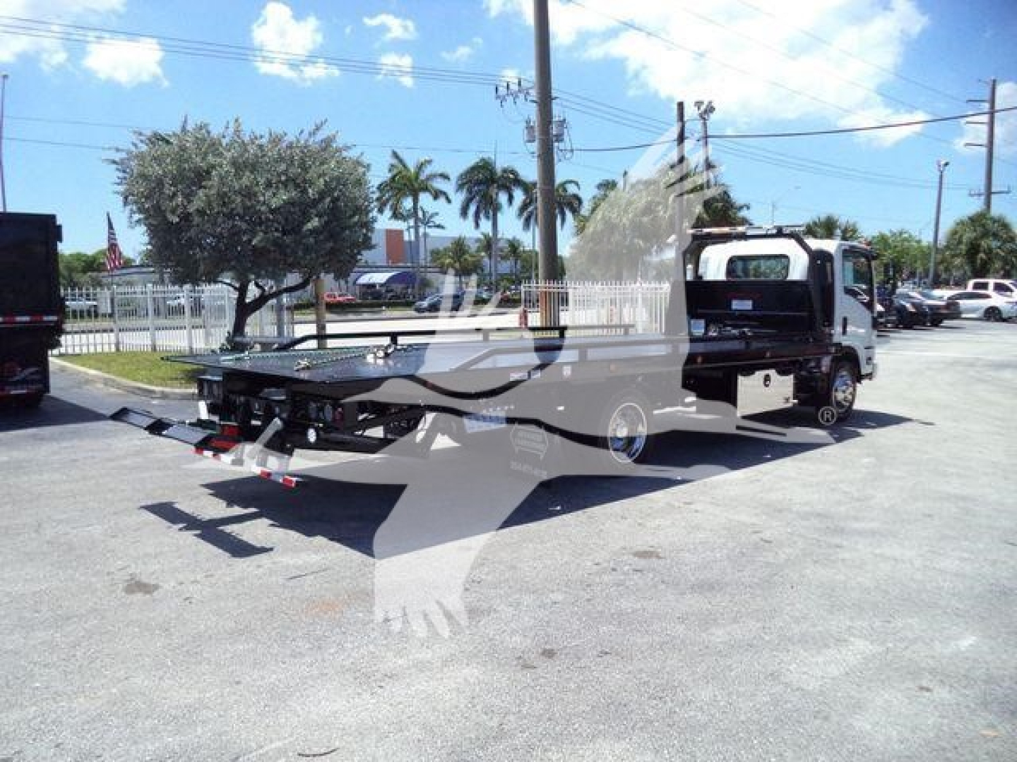 Isuzu NPRR flatbed rollback tow truck in a dealership lot, side view showing flatbed platform and towing equipment.
