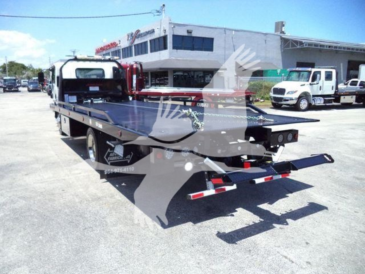 Isuzu rollback tow truck with flatbed in lot; visible text on side includes 'LONE STAR', 'LOADED' logos and contact numbers; background shows dealership building and other trucks.