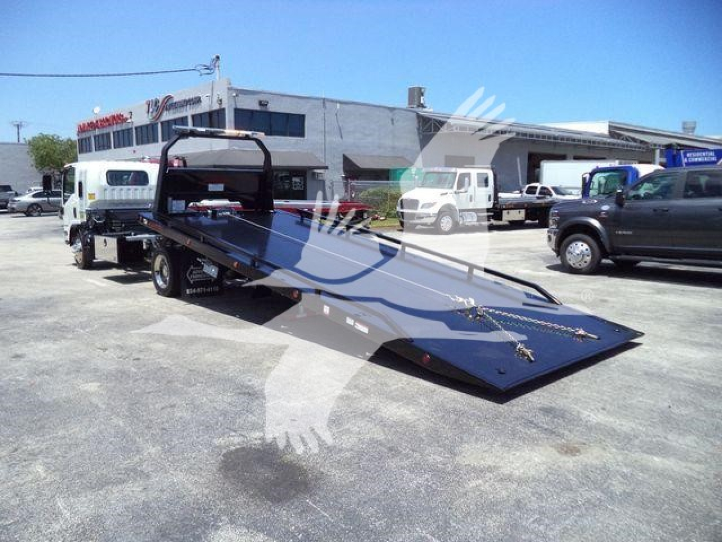Isuzu NPRR rollback truck with a long blue flatbed tilt bed in a dealership lot, several other trucks visible in background.