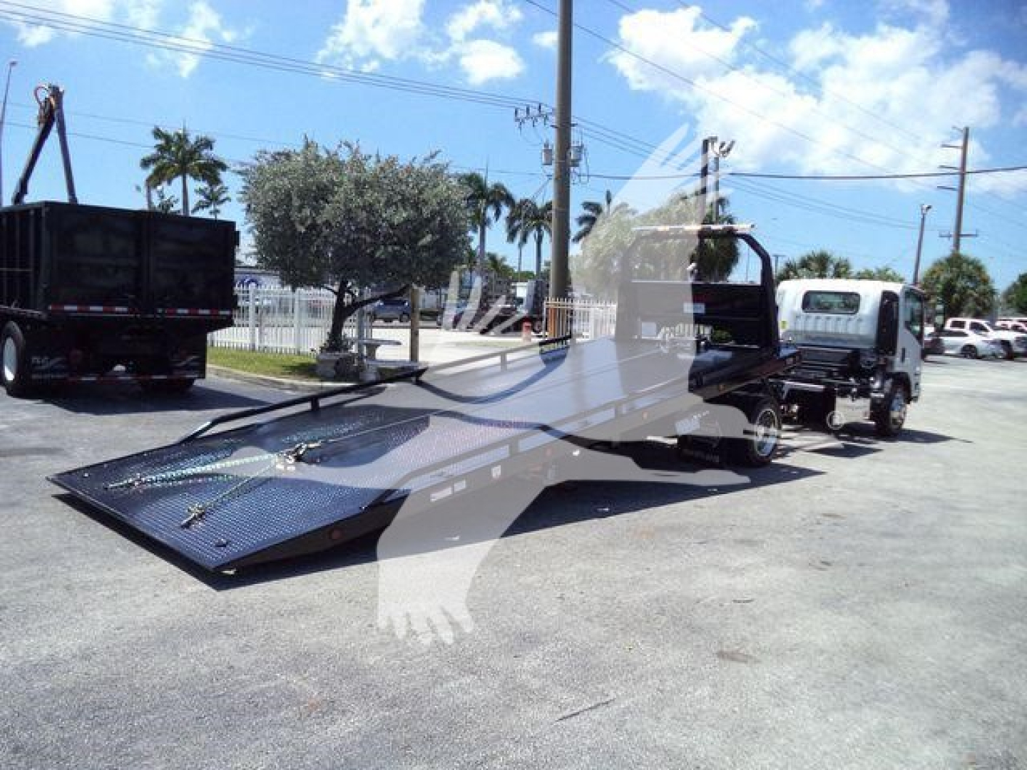 Isuzu NPRR truck rollback w/ flatbed and tow truck in lot, side view showing loading ramp and wheel lifts.