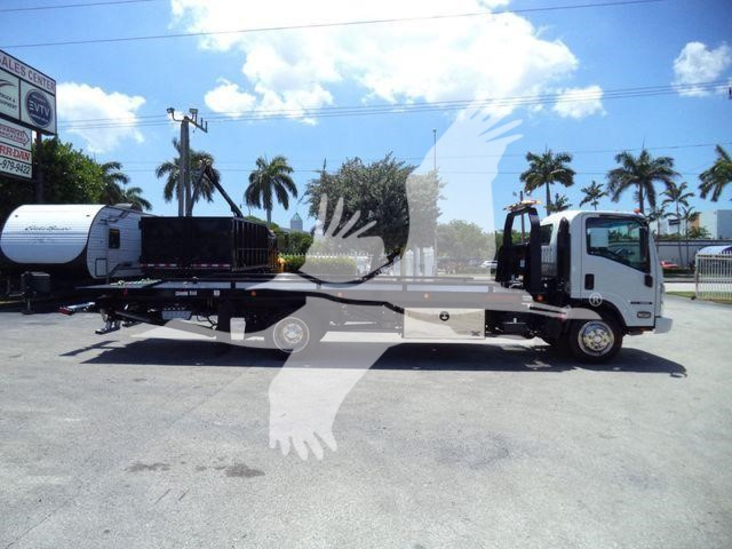 Isuzu NPRR rollback tow truck side view with flatbed and cab; palm trees and dealership lot in background.
