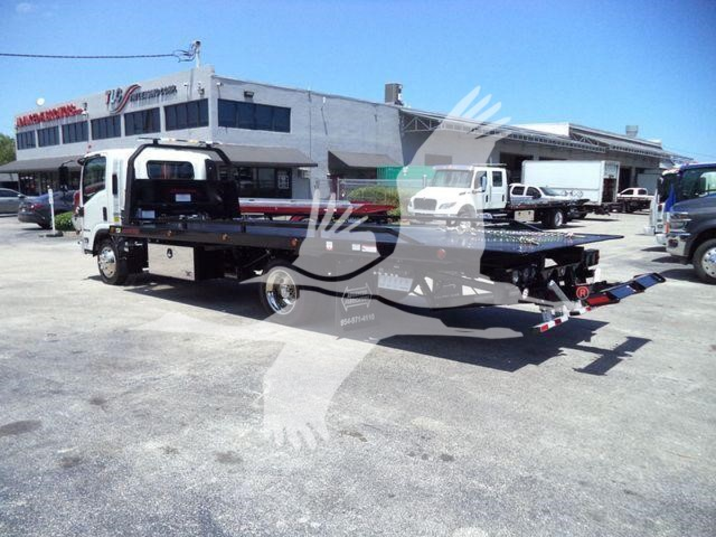 Isuzu NPR/NRR rollback tow truck with a flatbed and branding on building in background.