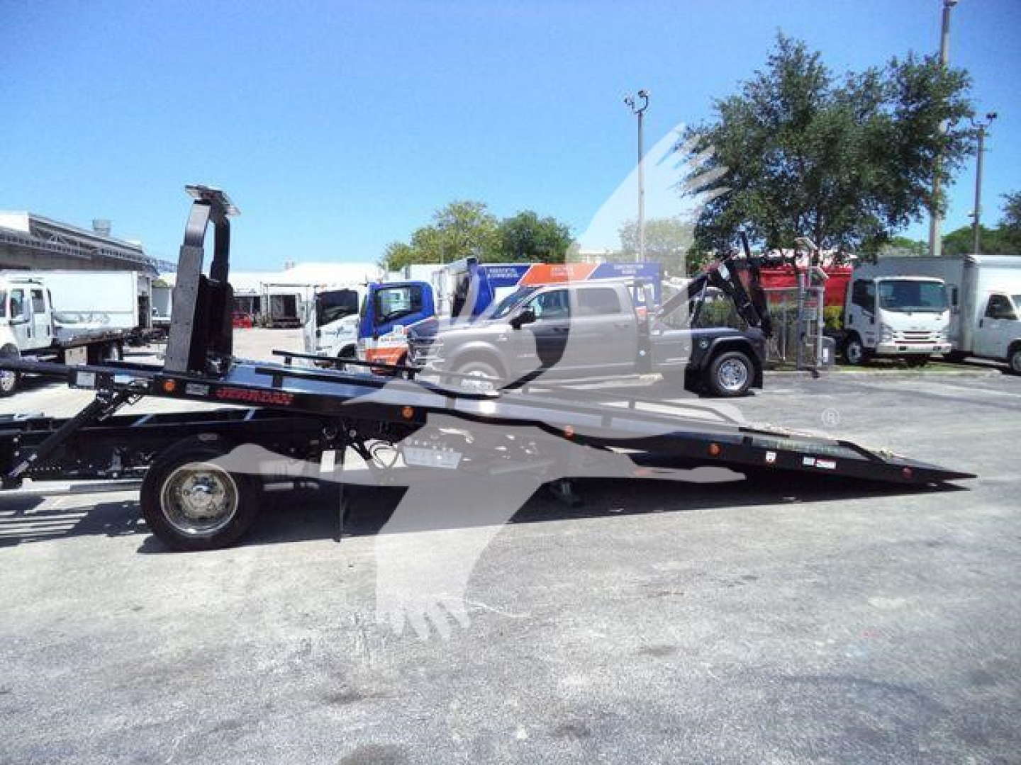 Isuzu rollback truck with metal flatbed tow bed and hydraulic ramps; visible sticker reads 'Isuzu' and there are other equipment and vehicles in background.