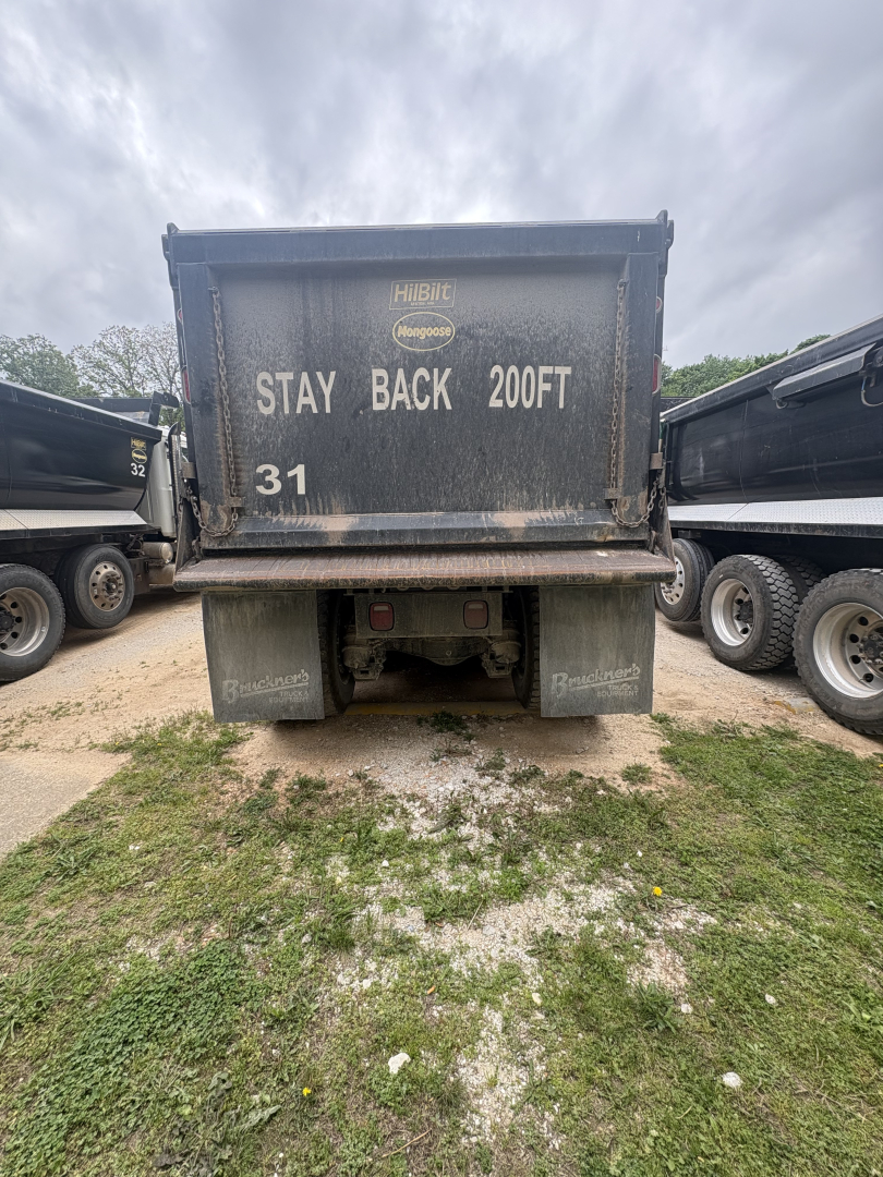 Rear view of a Mack dump truck with a heavy-duty dump bed. Text on bed reads: STAY BACK 200FT; 31. The area shows dirty bed and bumper with mud.