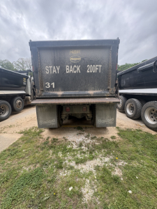 Rear view of a Mack dump truck with a heavy-duty dump bed. Text on bed reads: STAY BACK 200FT; 31. The area shows dirty bed and bumper with mud.