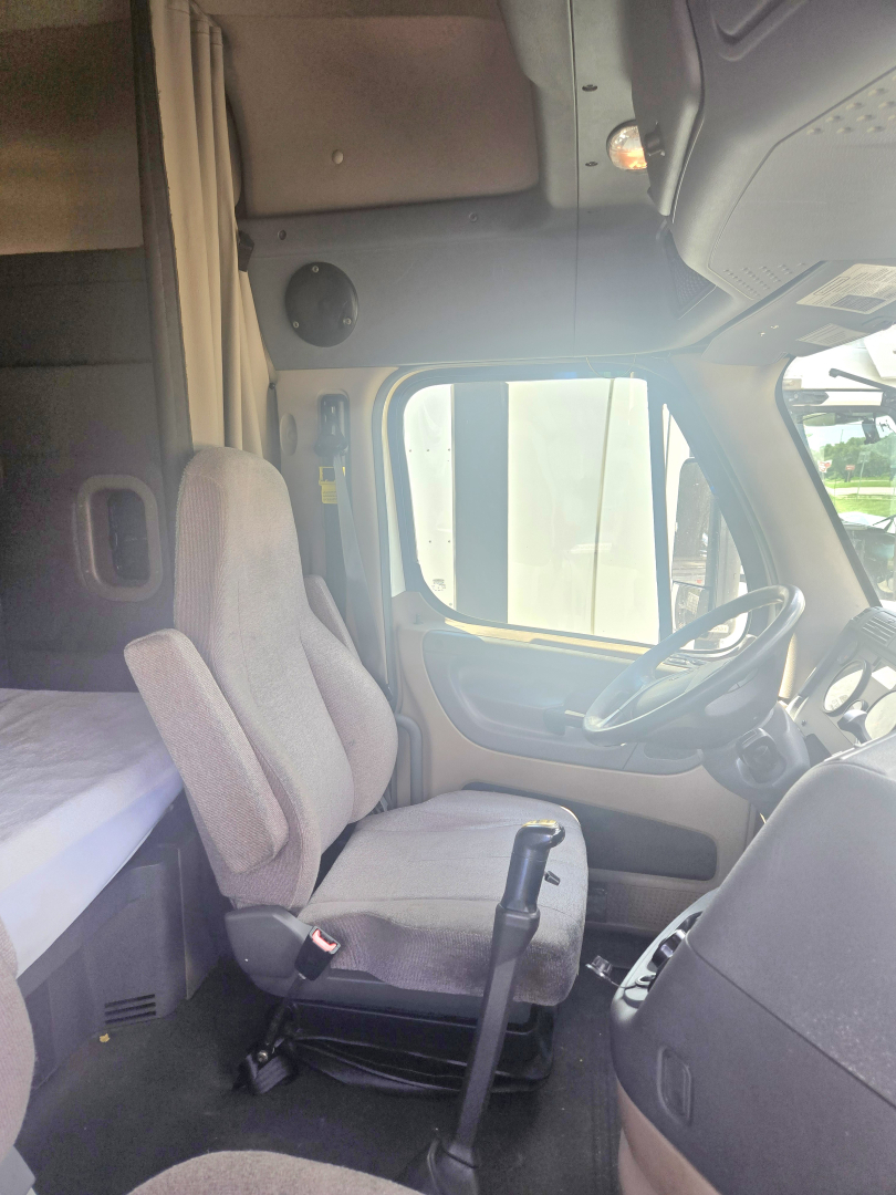 Truck cab interior of a 2019 Freightliner Cascadia midroof showing driver seat and dashboard view from passenger side, with sleeping area behind and bed.