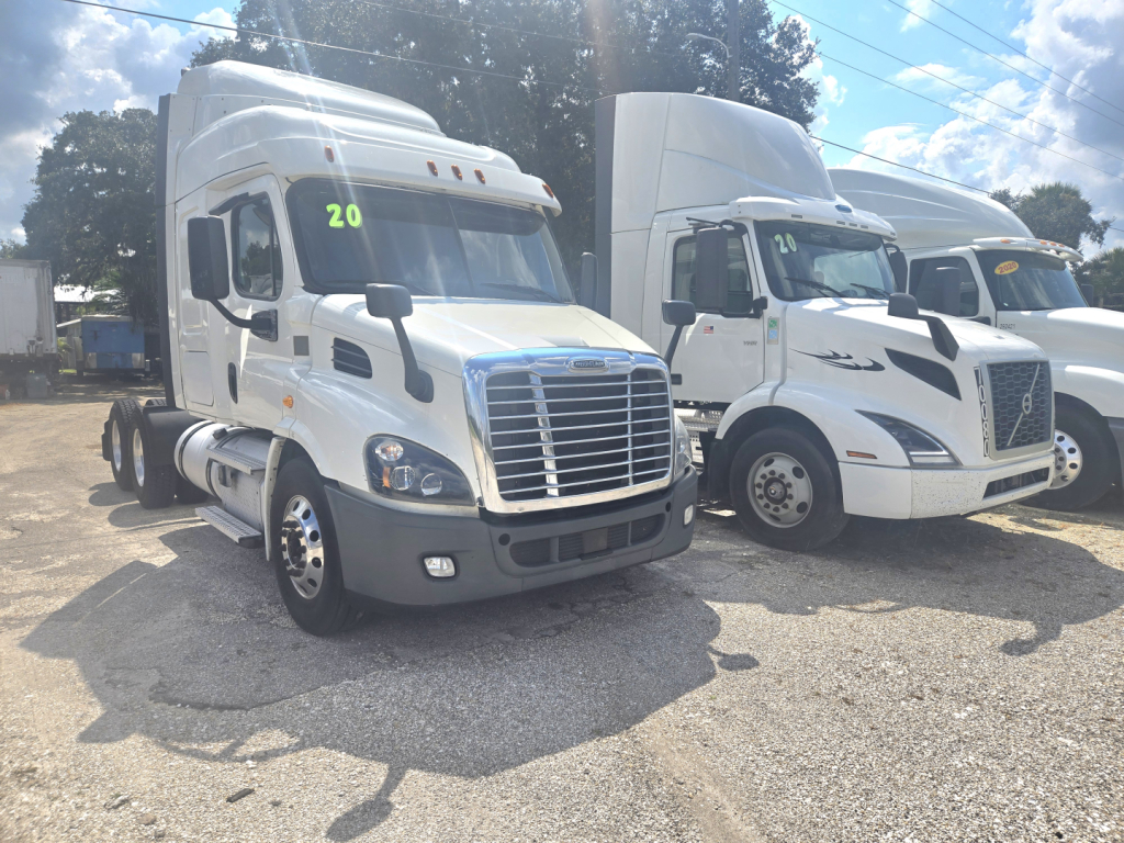 Truck cab showing Freightliner Cascadia midroof in a used-truck lot; white sleeper cab with side mirrors, grille, and front bumper visible; number 20 on windshield.