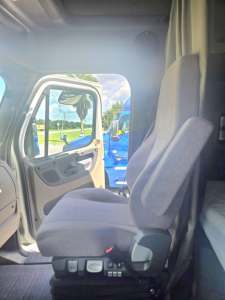 Truck cab interior showing driver seat and door with window, controls on seat base, and view into cab of a Freightliner Cascadia midroof.