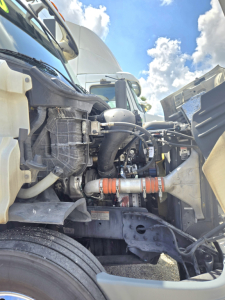 Truck engine bay of a 2019 Freightliner Cascadia midroof showing coolant hoses, turbo pipe, and air intake between bumper and cab.
