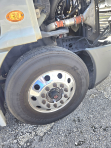 Fleet truck wheel and front suspension area of a Freightliner Cascadia midroof showing front tire, hub, and engine bay components with hoses and clamps.