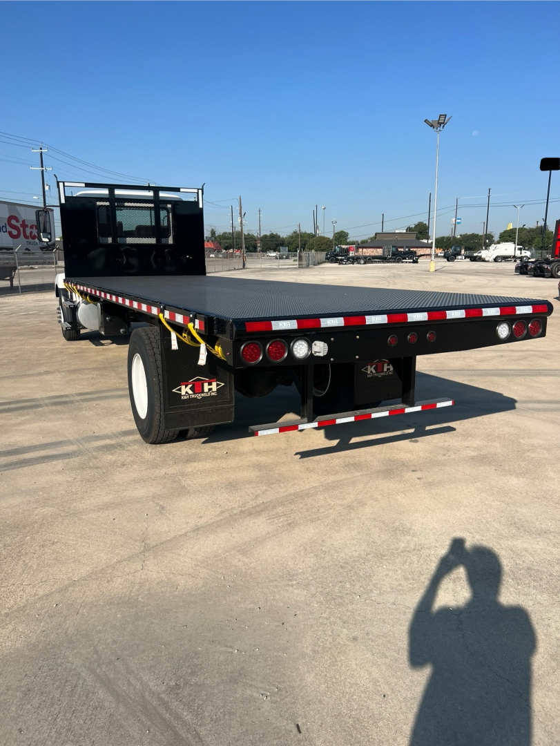 Flatbed truck rear view showing a flatbed with black rear guard, red and white reflective tape, round tail lights, bumper, and STK/KHP logo wheel fender. Clear blue sky.