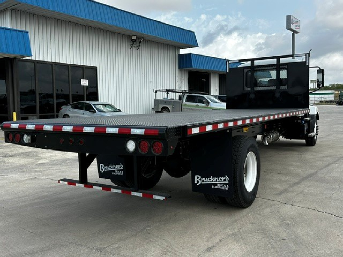 Flatbed truck chassis showing rear view of a 2026 Mack MD6 with a long flatbed, dual rear wheels, and Bruckner’s mud flaps.