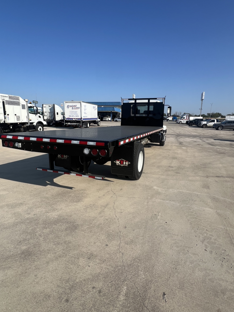 Flatbed trailer A-frame with black deck and red/white reflective edge, viewed from rear-left, Mack MD6 yard with trucks in background.