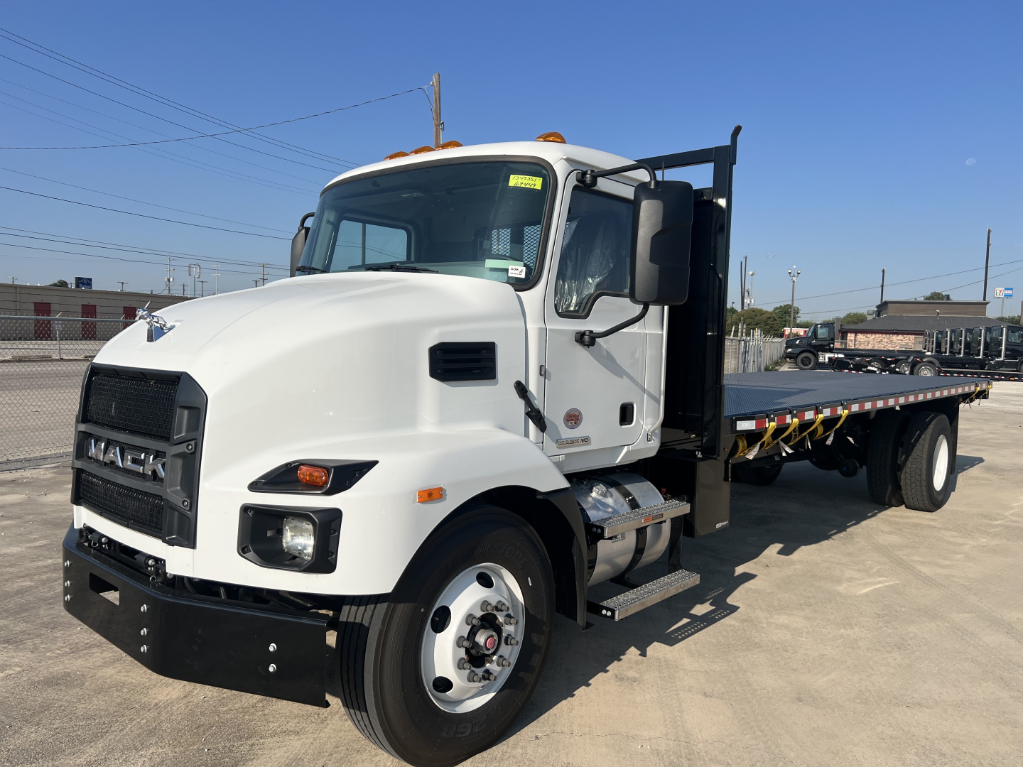 White Mack MD6 flatbed truck side view showing long flatbed with black rear chassis, dual stacked exhaust, and cab doors; VIN and stock number not visible in text on image.