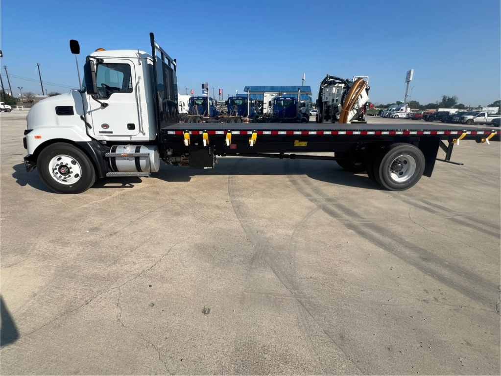 Flatbed truck side view of a 2026 Mack MD6 with long flatbed, white cab, black tires, and safety rails along the bed.