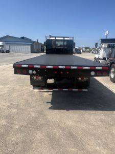 Flatbed truck bed of a 2026 Mack MD6 seen from rear with black deck and red reflectors. License plate area and rear underrun bar visible.