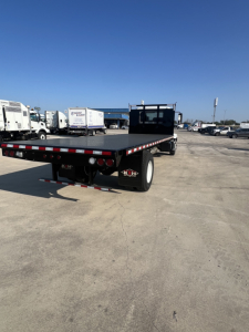Flatbed trailer A-frame with black deck and red/white reflective edge, viewed from rear-left, Mack MD6 yard with trucks in background.