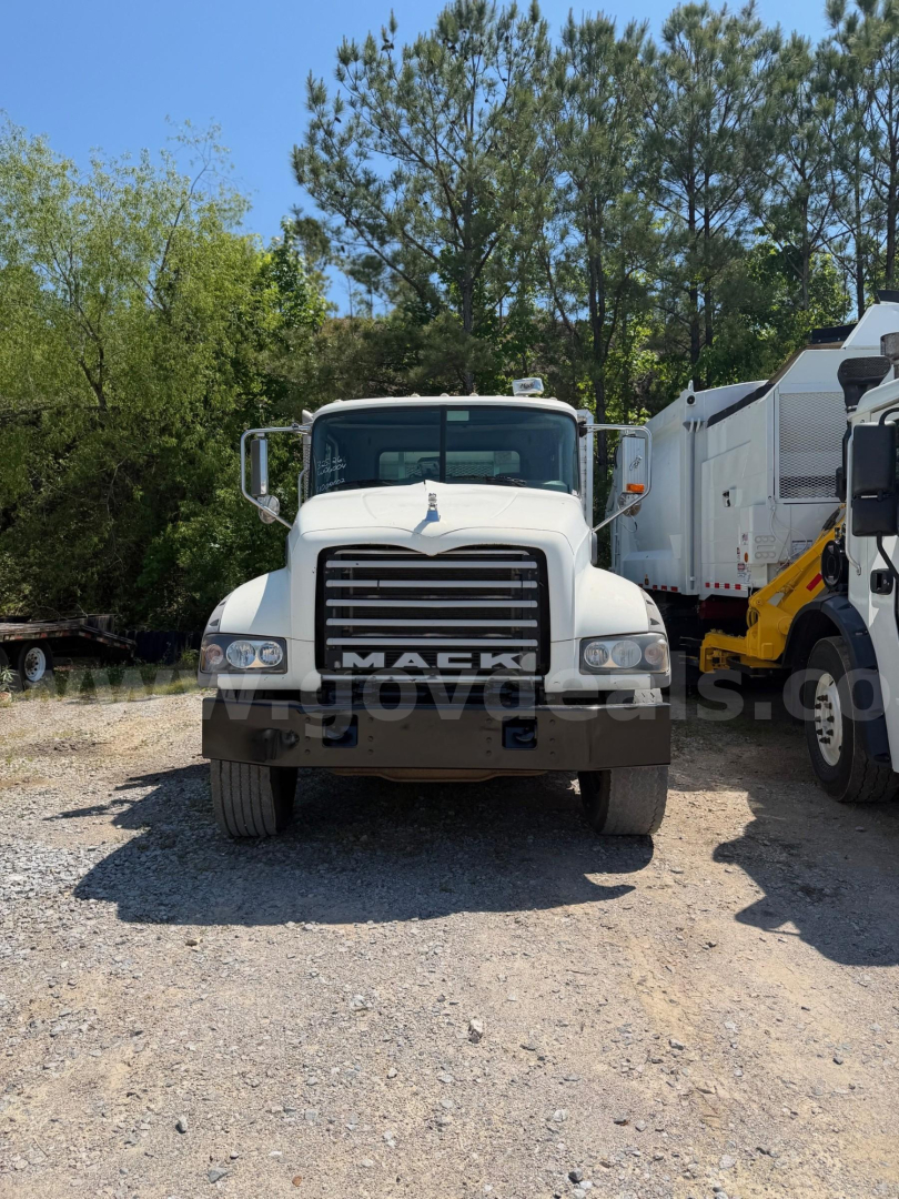 Front view of a white 2013 Mack Granite flatbed truck with large grille and bumper; visible text includes MACK on the grille and some reflective branding across the bumper.