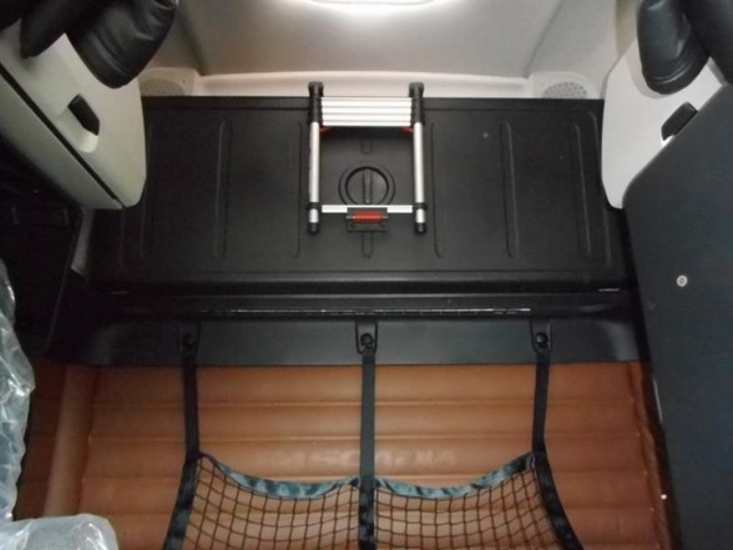 Truck cab interior showing under-seat storage area with brown quilted floor mat and rear storage hatch in Freightliner Cascadia 126 cabin.