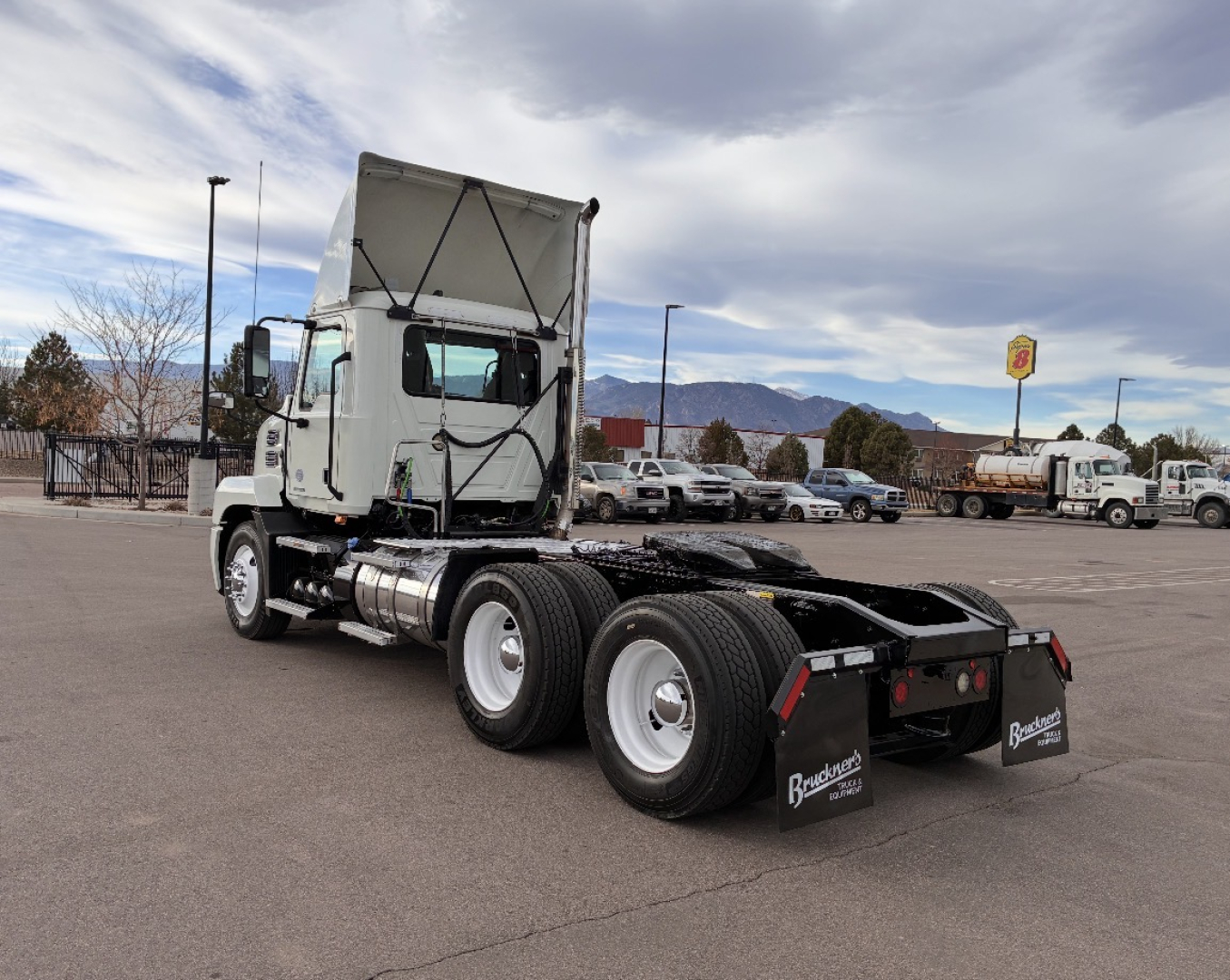 Truck chassis with raised cab on a daycab tractor showing rear chassis, tandem axles, fuel tanks, and cab in front of a parking lot.
