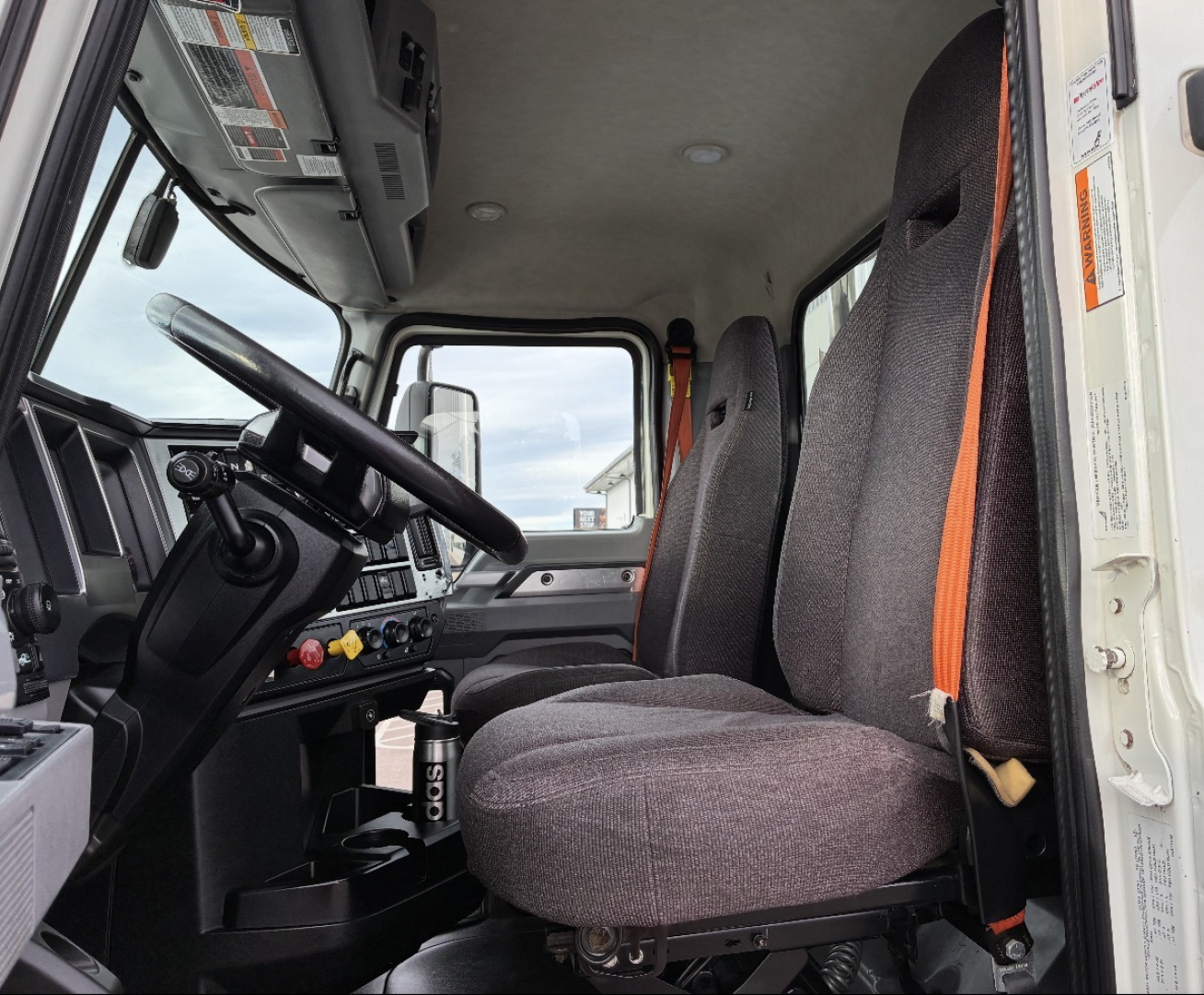 Truck cab interior: driver and passenger seats with grey fabric, orange seat belts, steering wheel, dashboard controls, and a view through the door to outside.