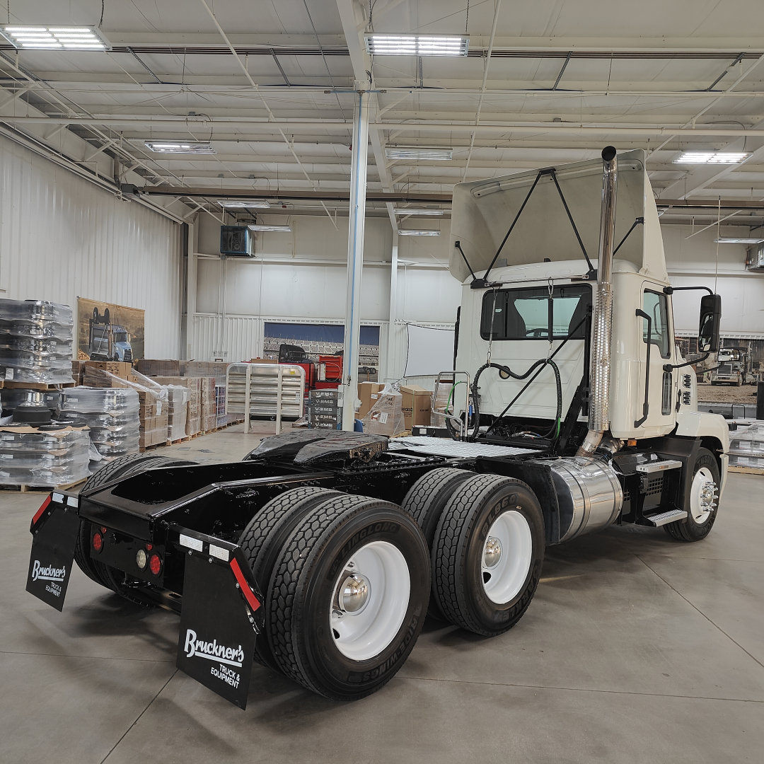 Mack AN64T daycab tractor in a warehouse, rear view showing dual rear axles, fifth-wheel missing, fuel tanks and exhaust stack visible.