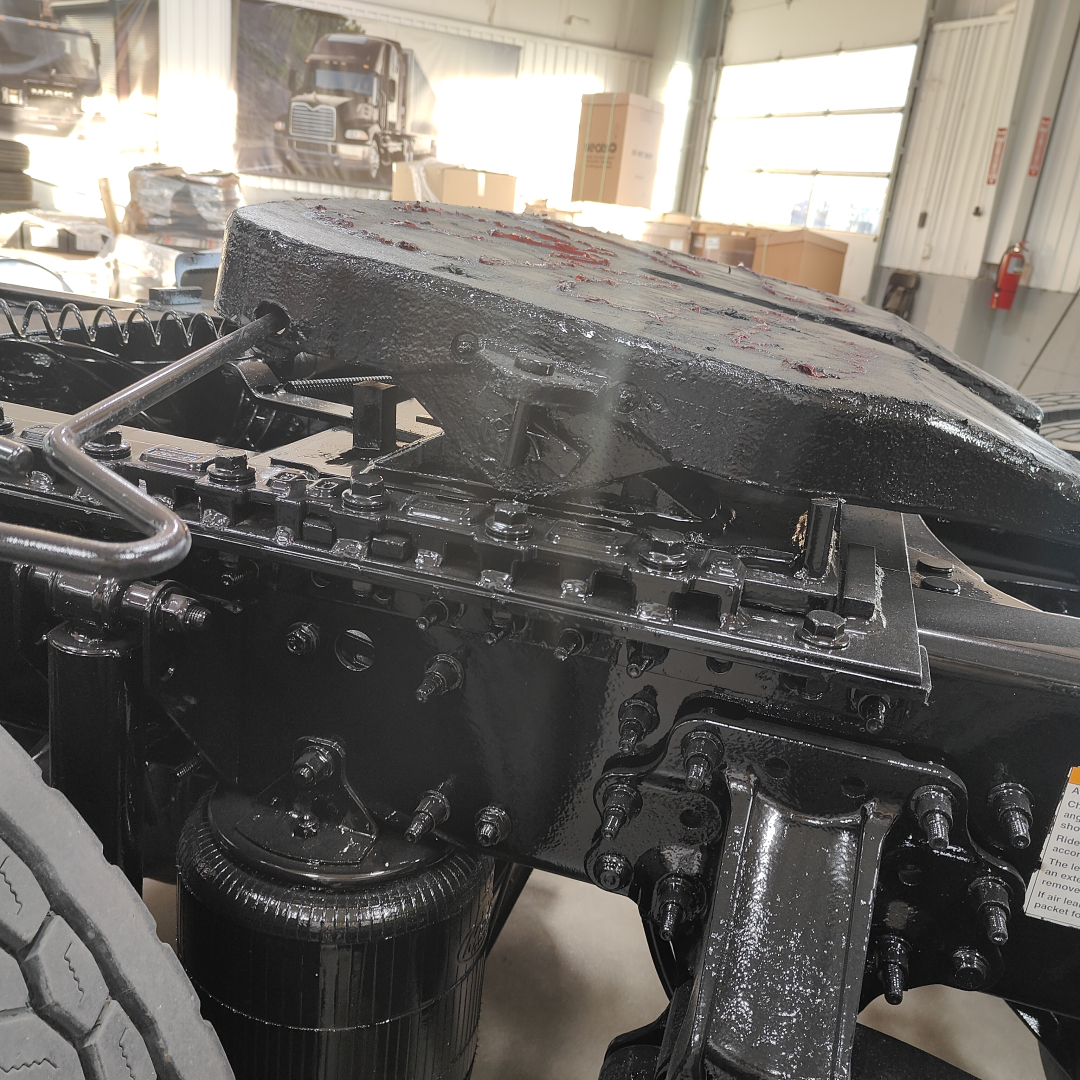 Truck engine bay and chassis area showing a black-painted component with hydraulic lines and a large block, in a well-lit shop. Visible text on sticker and metal fittings.