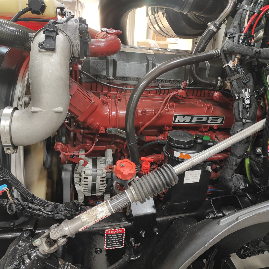 Truck engine bay showing red MEP-branded diesel engine with hoses, alternator, radiator hose, and coolant reservoir; visible labels, hoses, and wiring in a Mack AN64T cab.