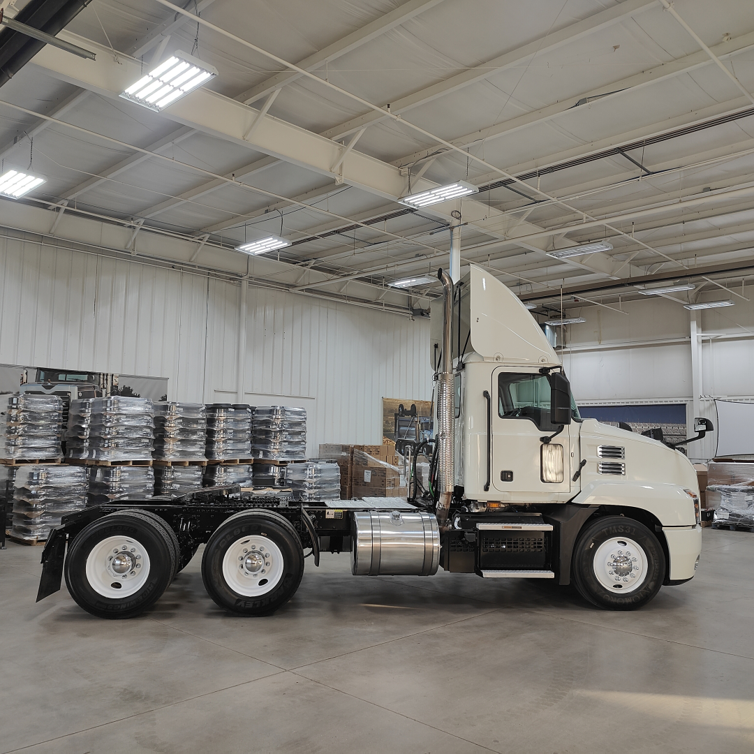 Truck side view of a 2020 Mack AN64T conventional daycab in a showroom, showing cab, sleeper fairing, tandem rear axles, and fuel tanks.