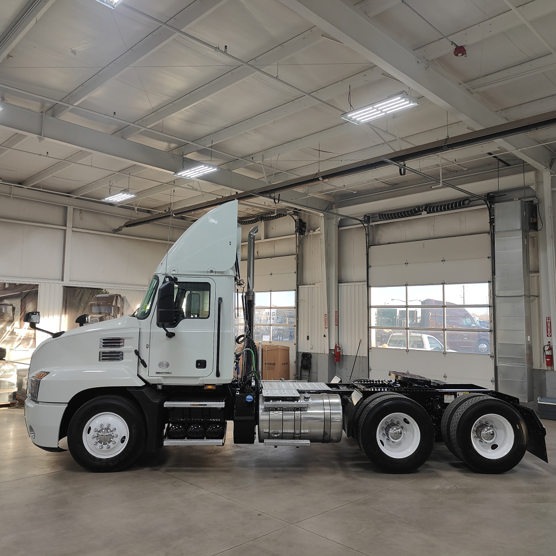 Truck cab and chassis profile inside showroom; white Mack AN64T with long nose, dual rear axle, fuel tanks, and cab steps visible.