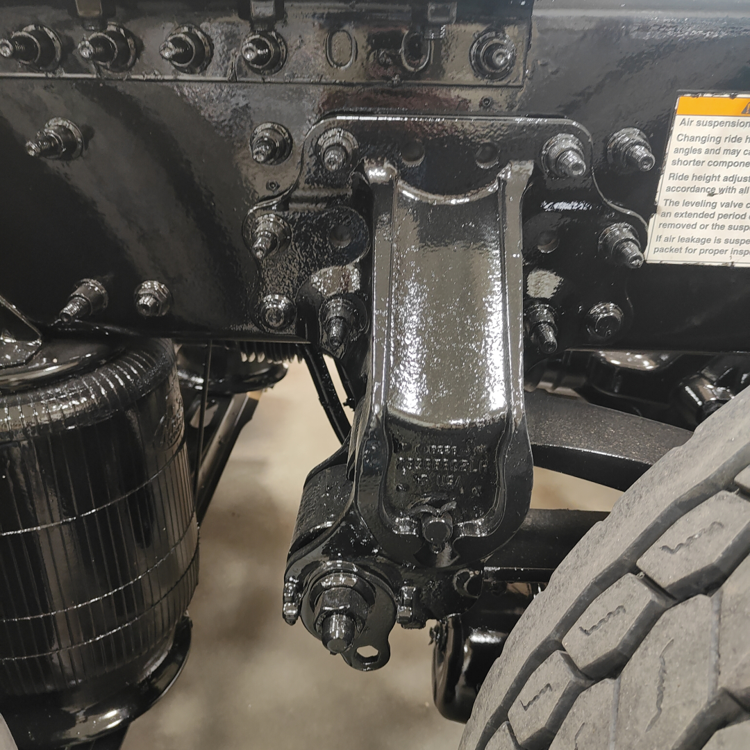 Component close-up underframe showing heavy-duty suspension linkage and bolts with black coating and grease; visible tire and protective label nearby.