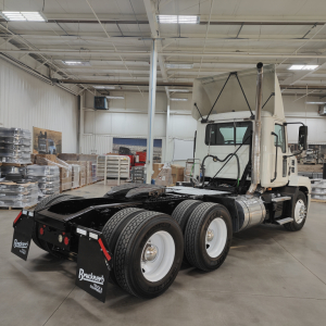 Mack AN64T daycab tractor in a warehouse, rear view showing dual rear axles, fifth-wheel missing, fuel tanks and exhaust stack visible.