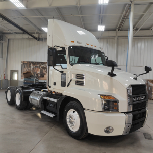 Mack AN64T tractor in a showroom setting, front three-quarter view, white cab with black accents, DEF and fuel tanks visible, no license plate.
