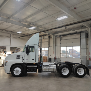 Truck cab and chassis profile inside showroom; white Mack AN64T with long nose, dual rear axle, fuel tanks, and cab steps visible.