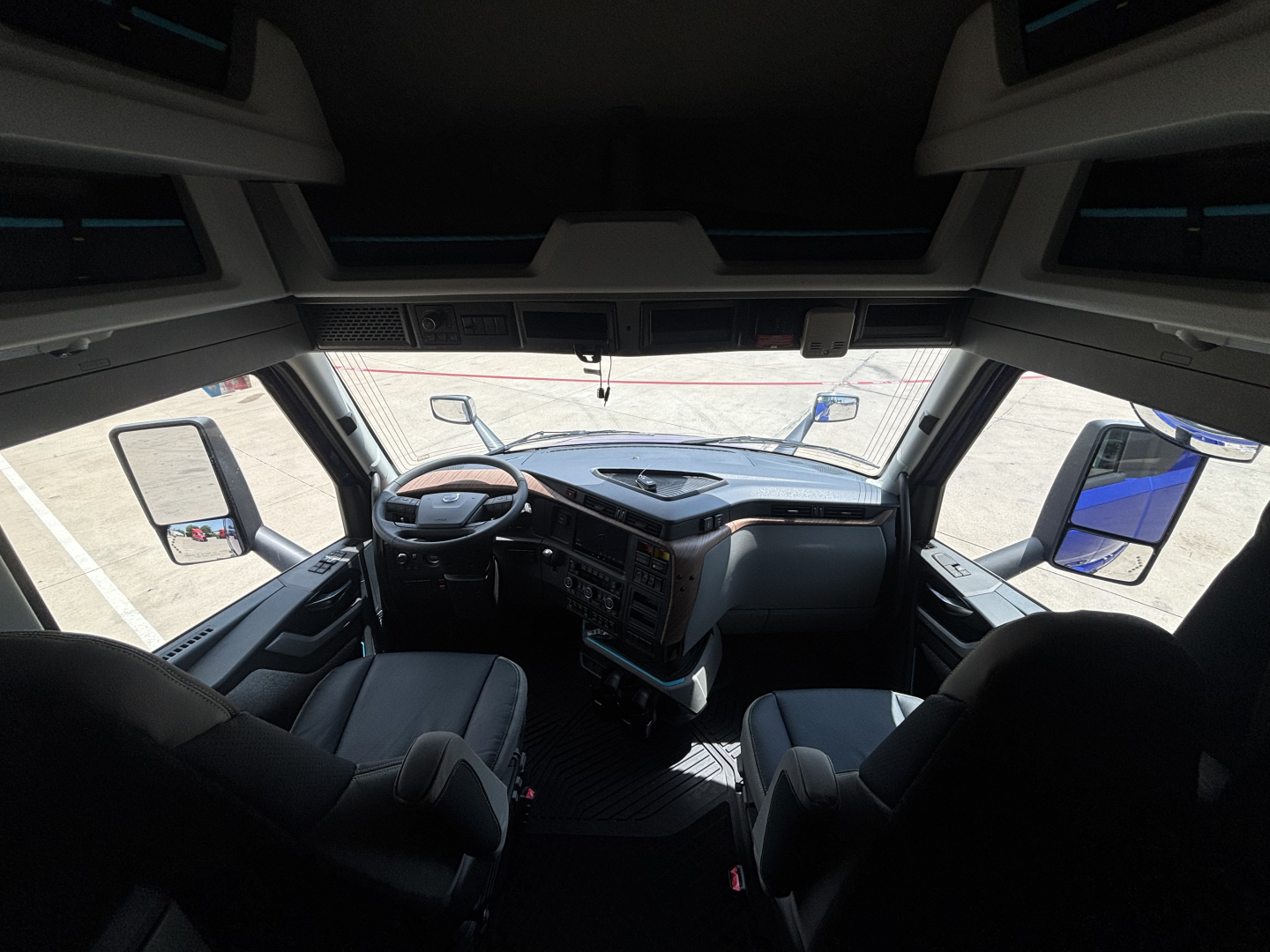 Truck cabin interior view from the sleeper cab, steering wheel, dashboard, driver and passenger seats, windshield, side mirrors.