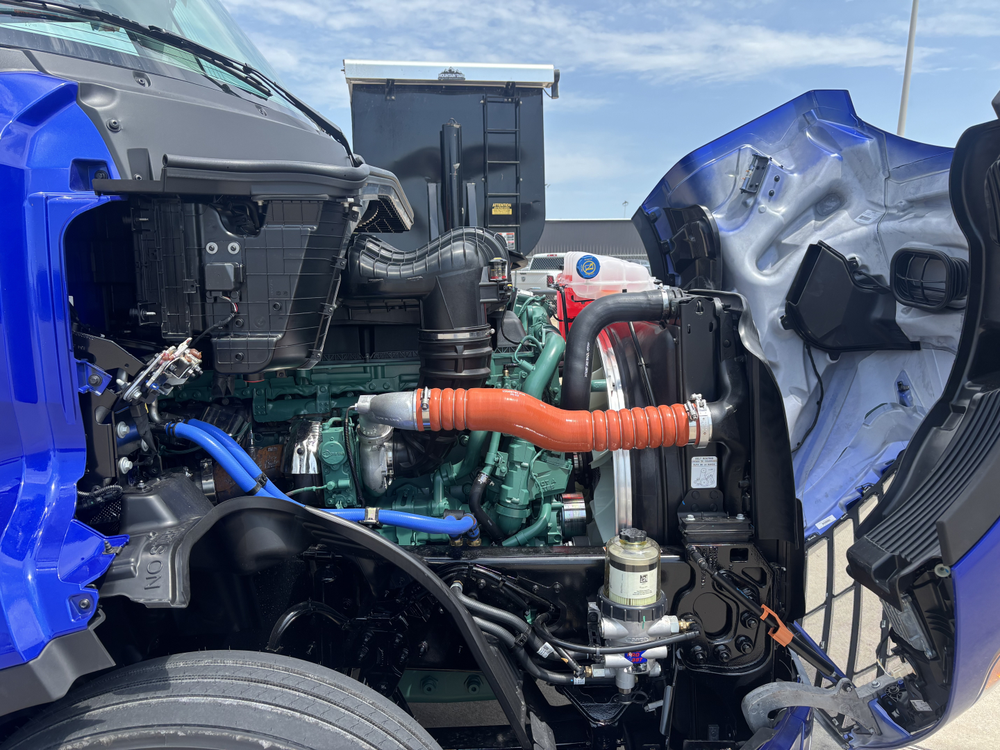 Volvo VNL64T860 engine bay view showing turbo hoses, coolant lines, and hydraulic components with blue cab exterior visible.