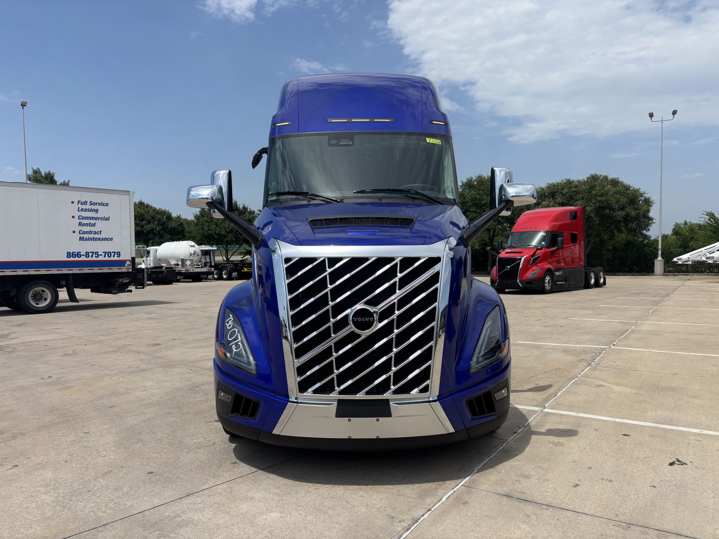 Front view of a blue 2027 Volvo VNL64T860 tractor in a lot, chrome grille with diagonal bars, large bumper, and side mirrors visible.