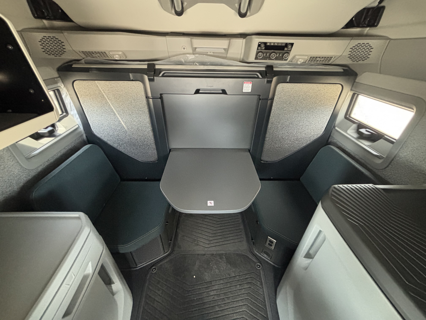 Interior cabin of a 2027 Volvo VNL64T860 sleeper showing front table/dinette area with fold-down seats and compact TV shelf; overhead dash cluster at front.