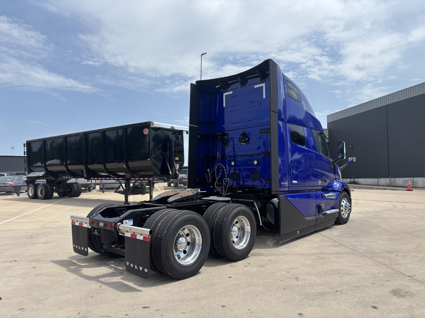 Volvo VNL tractor in blue cab with dual rear axles and fifth wheel, view from left-rear showing sleeper cab, trailer in background, clear sky.
