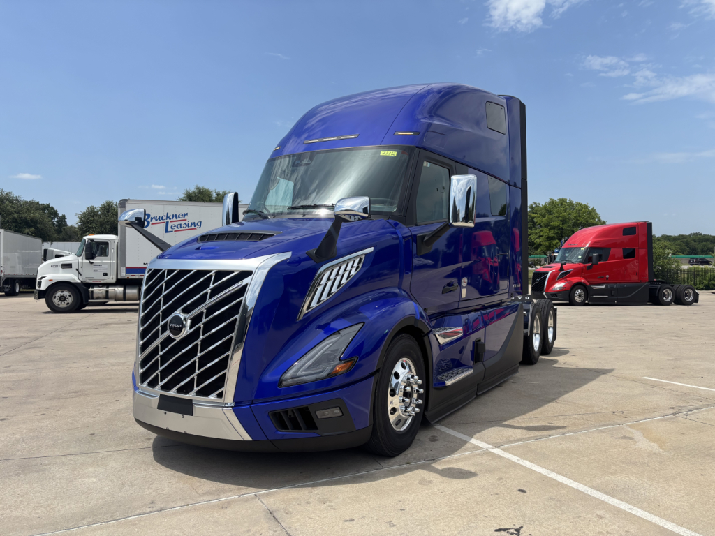 Blue Volvo VNL64T860 tractor-trailer in a dealership lot, front three-quarter view showing chrome grille, large aerodynamic mirrors, and sleeper cab.