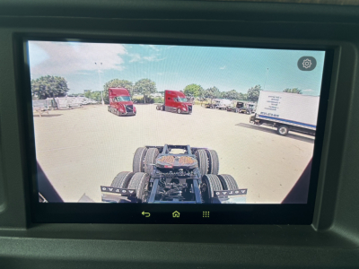 Truck cab view showing rear of Volvo VNL chassis with dual rear axles and parking lot with red trucks and a white trailer in background.