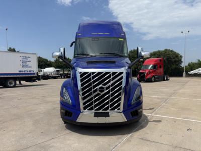 Front view of a blue 2027 Volvo VNL64T860 tractor in a lot, chrome grille with diagonal bars, large bumper, and side mirrors visible.