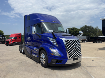 Blue Volvo VNL64T860 semi-truck in a lot, front three-quarter view, chrome grille, sleeper cab, other trucks in background.