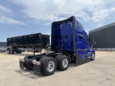 Volvo VNL tractor in blue cab with dual rear axles and fifth wheel, view from left-rear showing sleeper cab, trailer in background, clear sky.