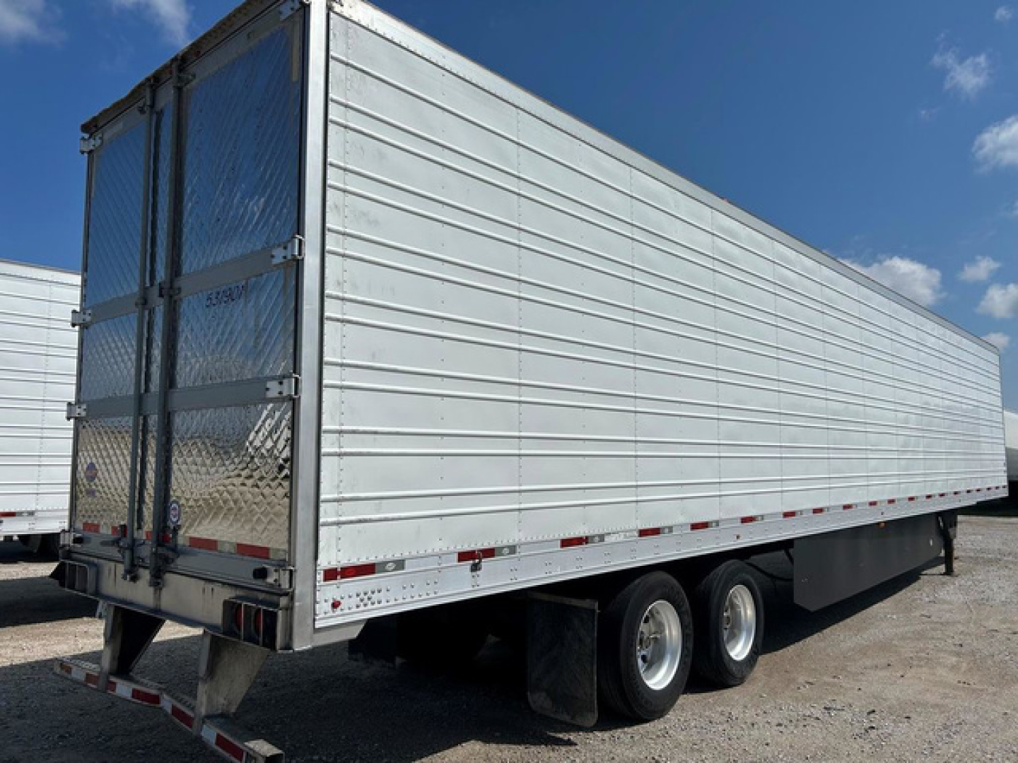 Utility reefer trailer with aluminum side panels, dual rear doors, and rear bumper; visible are taillights, reflective tape, and a support wheel area.