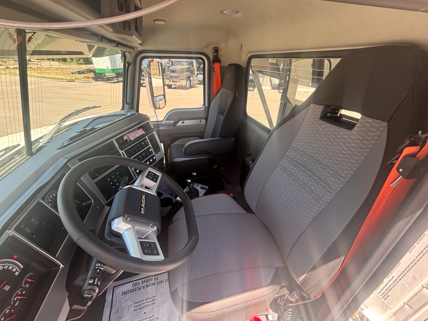 Interior of a 2026 Mack GR64B cab showing steering wheel, dashboard, driver's seat, and passenger seat with gray fabric and safety harness; view from open door.