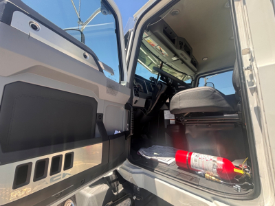 Cab interior of a 2026 Mack GR64B rolloff truck showing driver seat, steering wheel, open door, dashboard, and a red fire extinguisher on the floor.