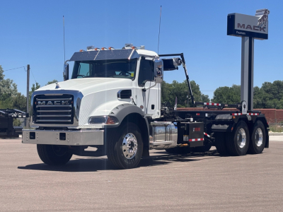 White Mack GR64B rolloff truck in a dealership lot showing front grille, large bumper, dual rear axles, and flatbed hooklift.