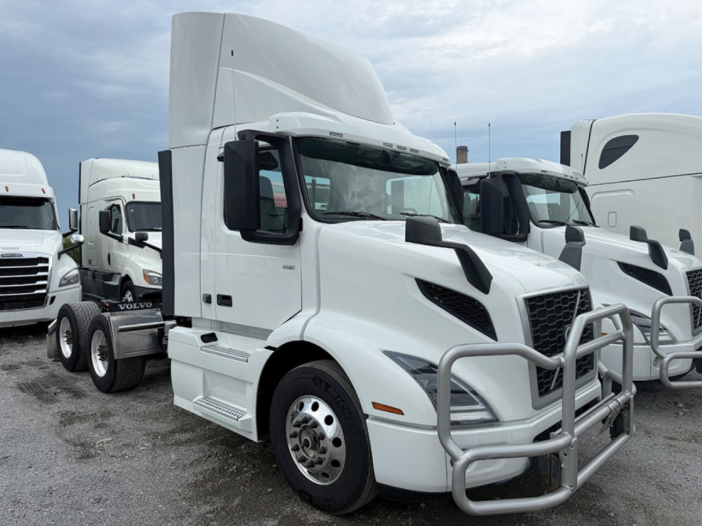 Volvo VNR 300 tractor-trailer cab in white, front grille guard, large sleeper, side steps, parked among other trucks.